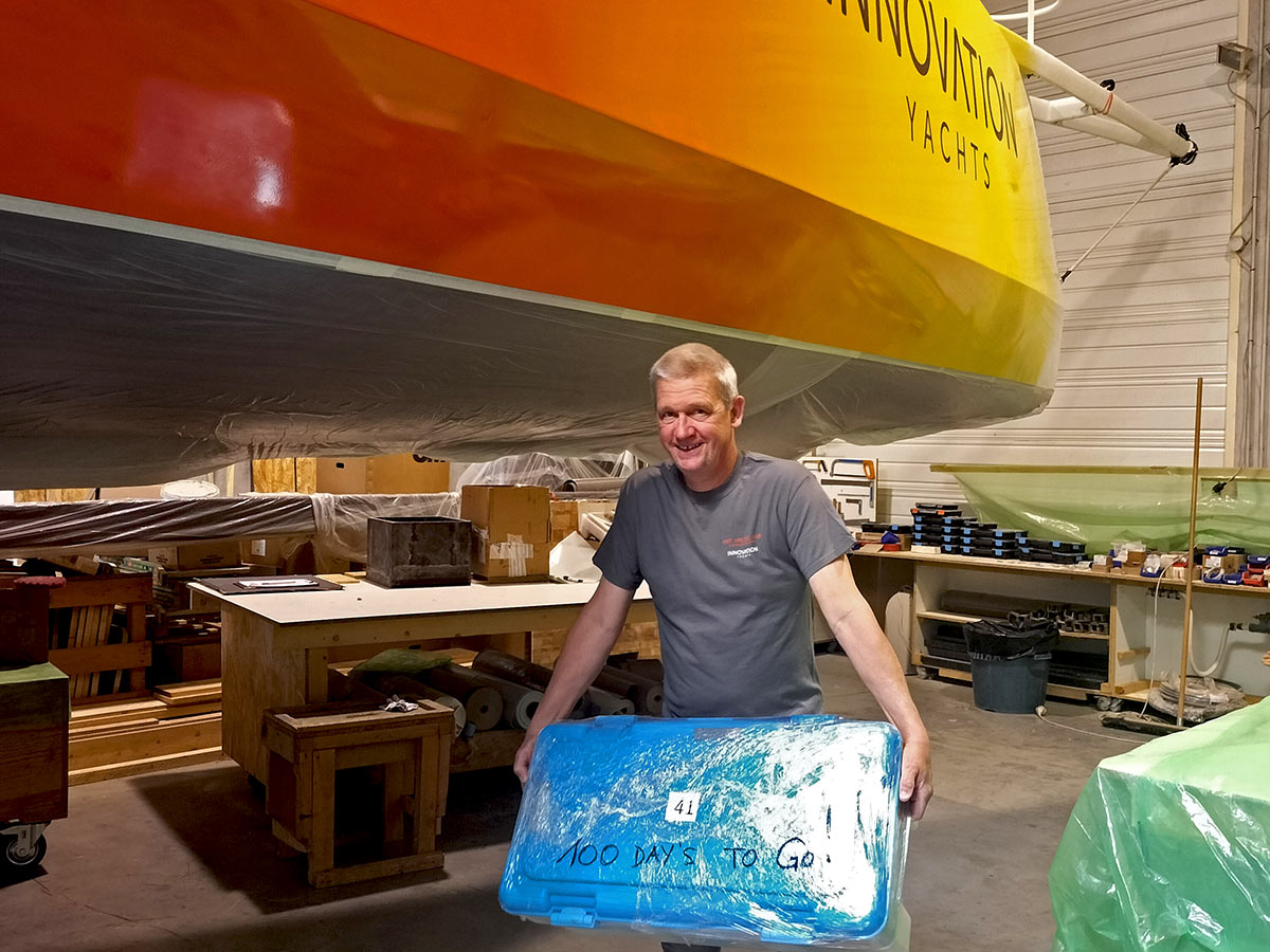 Norbert holding box in his hand, marked on it is "100 days to go!", standing in front of Open60AAL Innovation Yachts