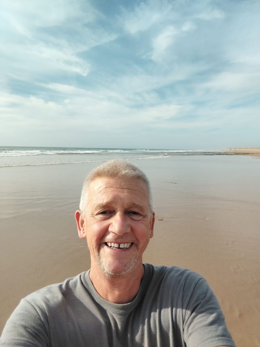 Norbert is smiling on the beach in front of the sea under a slightly cloudy sky with many shades of blue