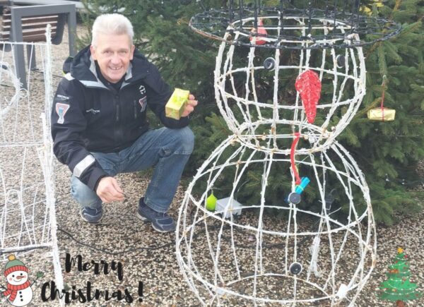 Norbert with an electric snowman on his left and a little gift in his hand in front of a Christmas tree in Vairé