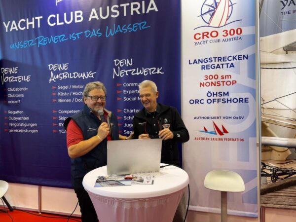 Norbert is standing near Gottfried Rieser at the YCA booth and he laughs while Gottfried is speaking in the microphone