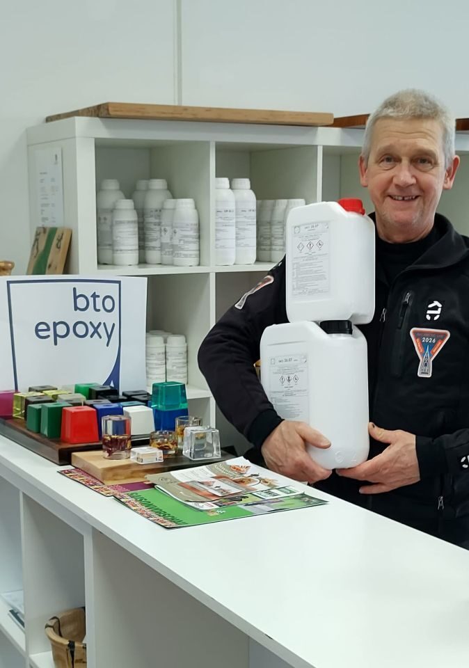 Norbert is standing behind the bto-epoxy counter, in front of shelves with samples, with two cans of resin and hardener stacked in his arms.