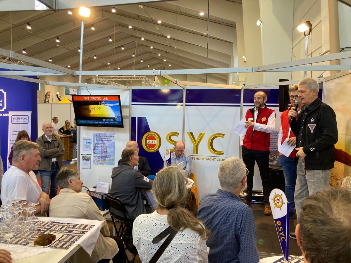 Norbert is speaking on the stage of OSYC booth at Austrian Boat Show with people carefully listening to him and a screen shows picture from the Open60AAL