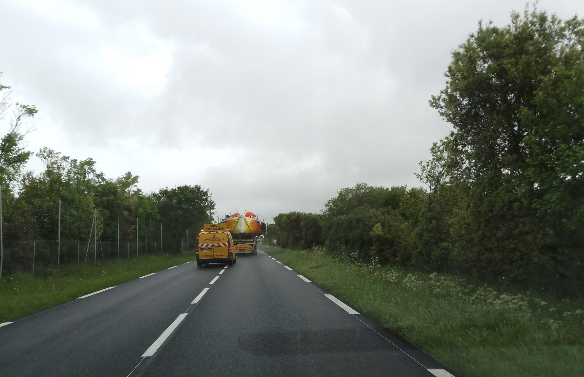 Auf einer Straße zwischen Bäumen und Hecken steht der Open60AAL auf einem Lkw, der Bug ist zu sehen, und ein kleiner Lieferwagen sorgt für die Sicherheit des Transports.