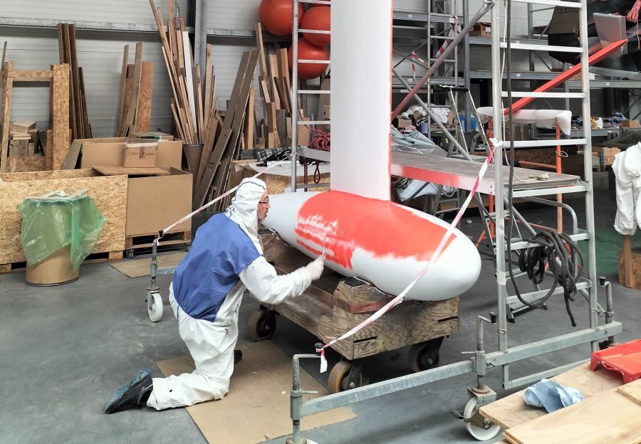 A team member, wearing an overal with a hood, is painting the bulb of the keel with fluo antifouling.