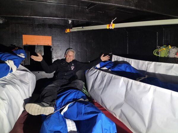 Norbert is sitting in the sails compartment, among the stored sails, opening his arm in a happy attitude