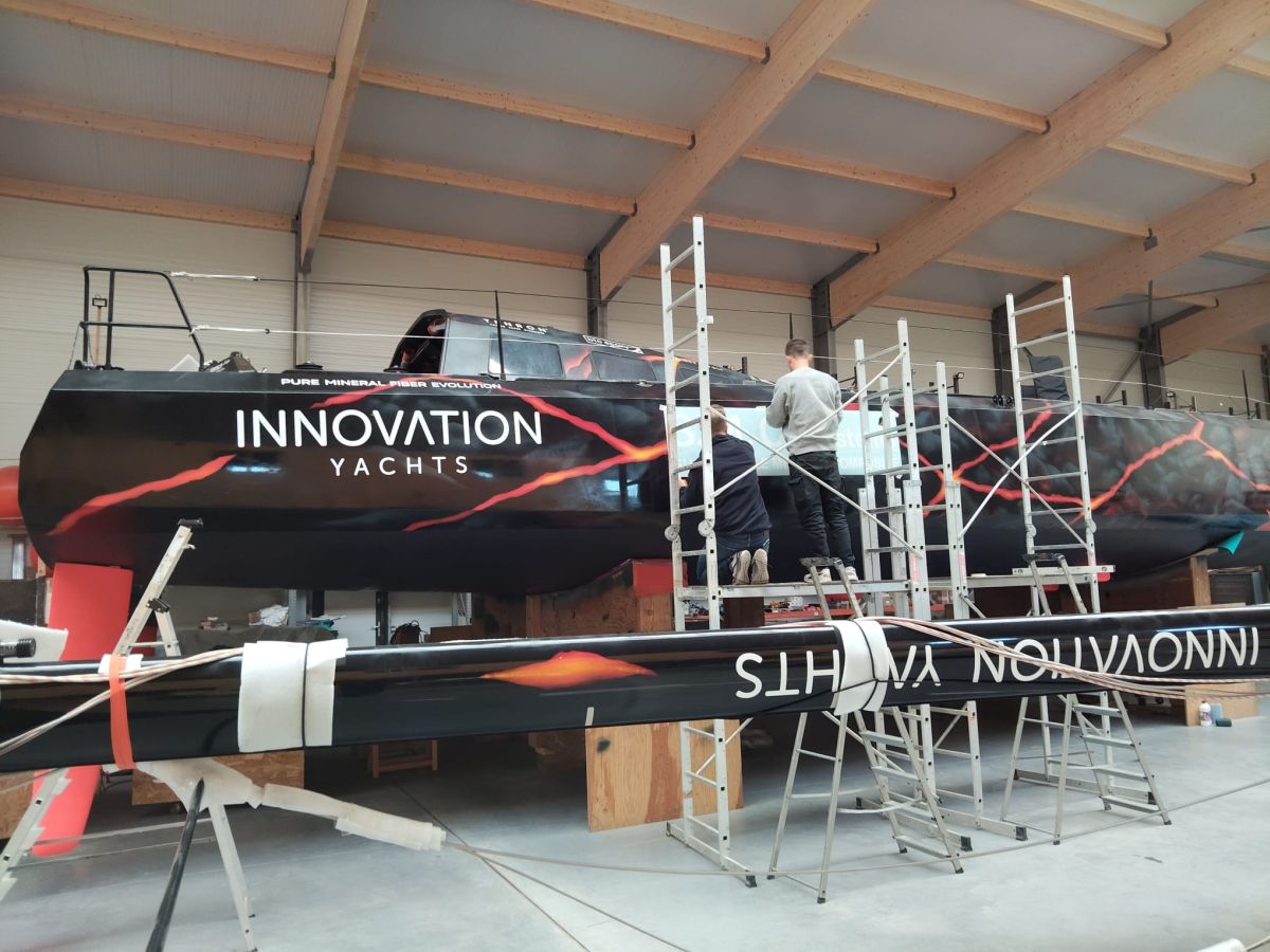 Two team members are posing a big sticker on the Open60AAL, in the shipyard.