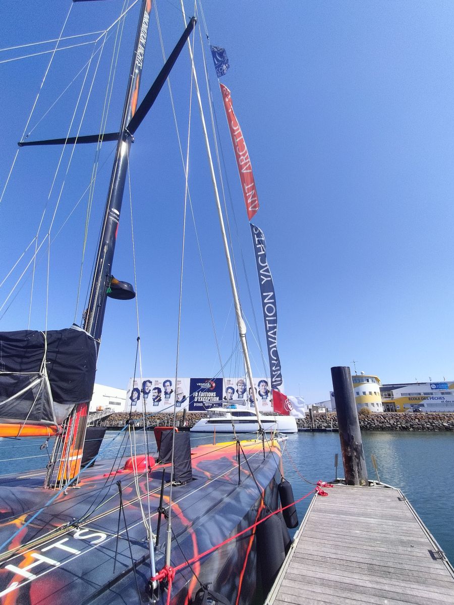 L'Open60AAL à Port Olona, avec ses drapeaux volants au vent devant les images en hommange aux vainqueurs du Vendée Globe